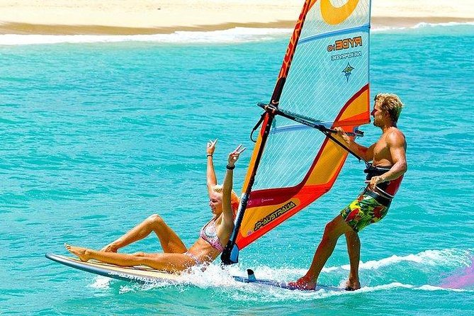 Two people on a windsurf board, one sitting and one operating the sail