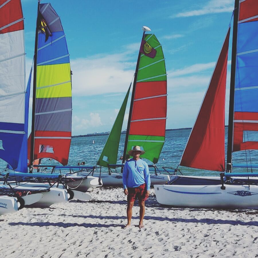 A person with a hat standing on a sandy beach surrounded by Hobie sailboats